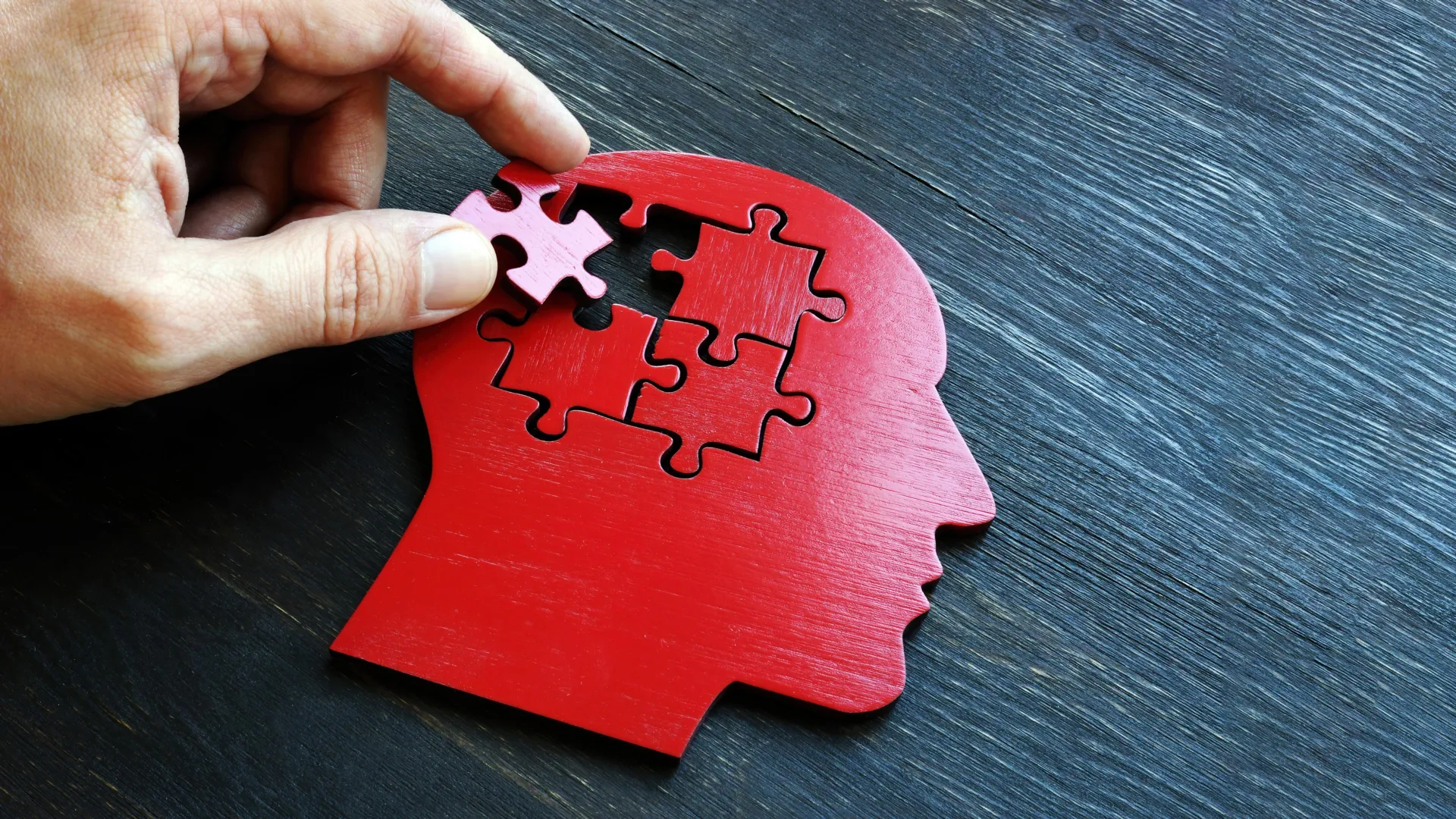 Hand placing a jigsaw puzzle piece into a red wooden head cutout representing brain health